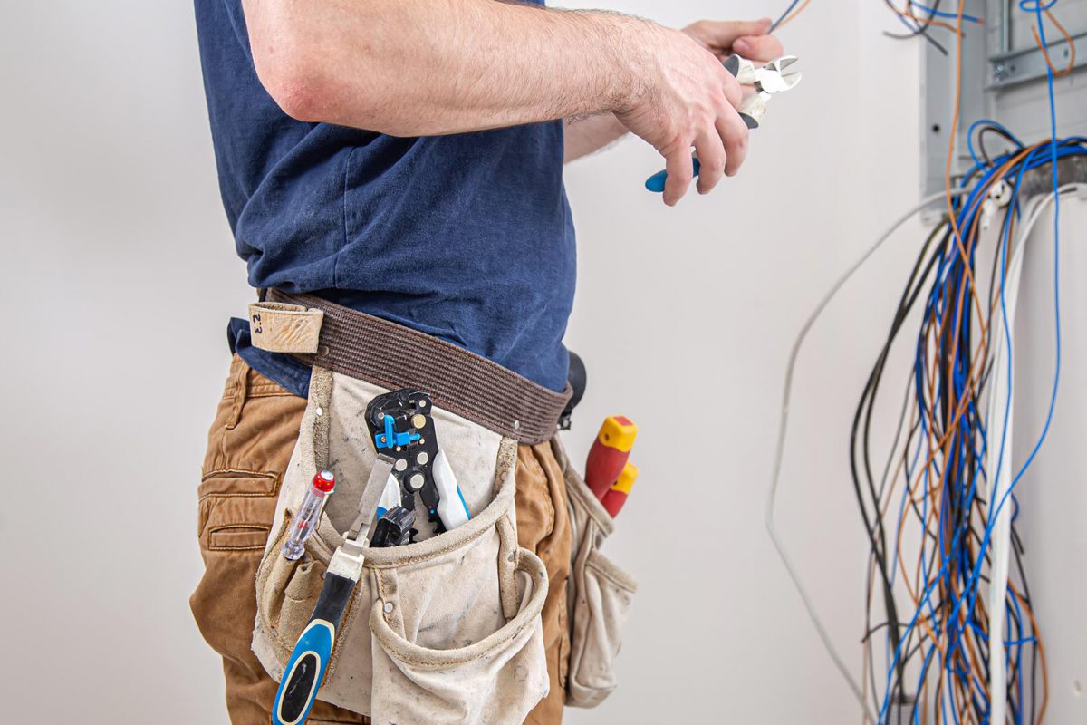 Electrician builder at work, examines the cable connection in the electrical line in the fuselage of an industrial switchboard. Professional in overalls with an electrician's tool.