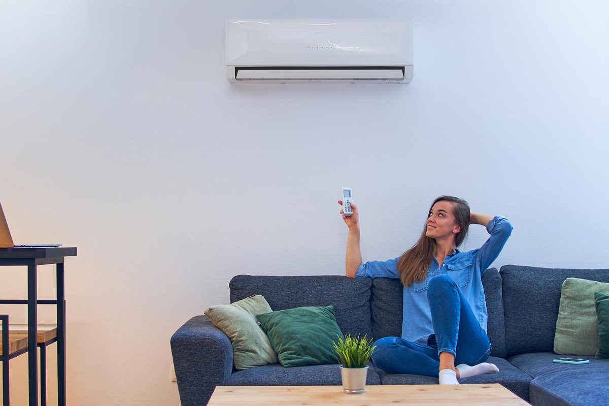 Young happy woman sitting on couch under air conditioner and adjusting comfort temperature with remote control at modern home